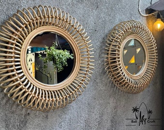 May include: Two round rattan mirrors with a sunburst design. The mirrors are hanging on a gray wall. The larger mirror reflects a plant hanging in front of it.