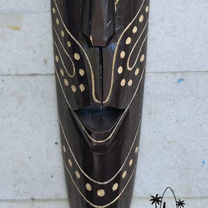 May include: A dark brown wooden mask with a carved face and white painted details. The mask has a wide mouth and a small nose. The mask is decorated with white dots and lines.