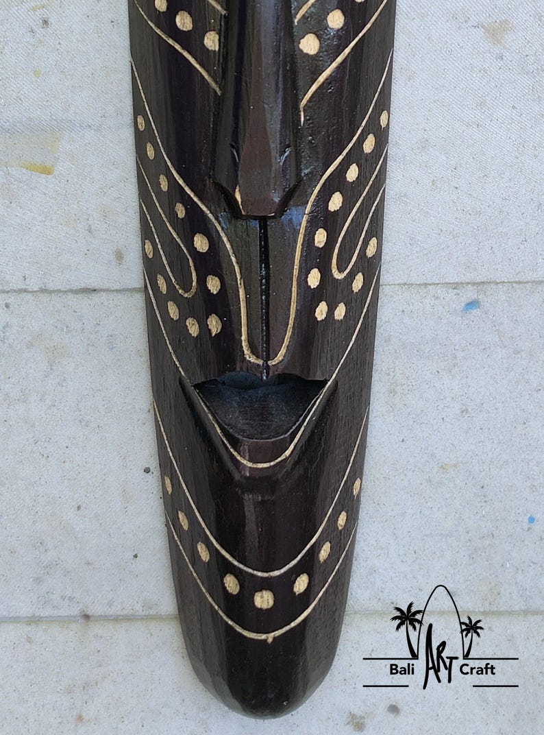 May include: A dark brown wooden mask with a carved face and white painted details. The mask has a wide mouth and a small nose. The mask is decorated with white dots and lines.