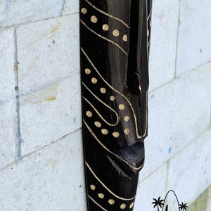 May include: Black wooden mask with a stylized face and gold dot pattern. The mask is hanging on a white brick wall. Bali Art Craft is printed below the mask.