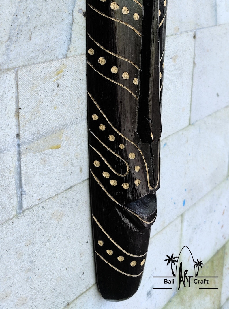 May include: Black wooden mask with a stylized face and gold dot pattern. The mask is hanging on a white brick wall. Bali Art Craft is printed below the mask.