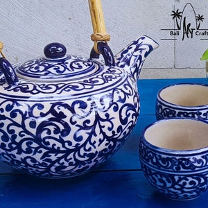 Op de afbeelding: Een witte theepot van keramiek met blauwe bloemmotieven en bamboe handvatten, samen met twee bijpassende theekopjes. De theepot heeft een deksel en een tuit. De theekopjes zijn rond en hebben een blauwe bloemmotief rond de rand.