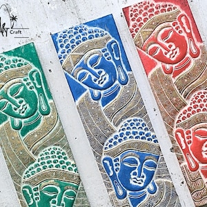 May include: Three wooden wall hangings with carved Buddha faces. The Buddha faces are in shades of blue, green, and red. The background of each carving is a gold color with intricate patterns.