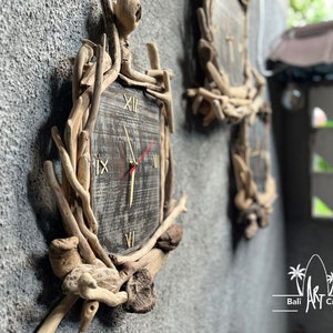 Op de afbeelding: Een rustieke houten klok met Romeinse cijfers op een verweerd houten gezicht. De klok is omgeven door takken van drijfhout en is gemonteerd op een grijze muur.