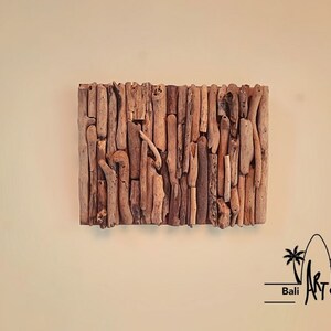 May include: A wall art piece made from driftwood, arranged in a vertical pattern. The wood is a light brown color and has a natural, rustic look. The piece is mounted on a white wall. The text "Bali Art Craft" is visible in the bottom right corner of the image.