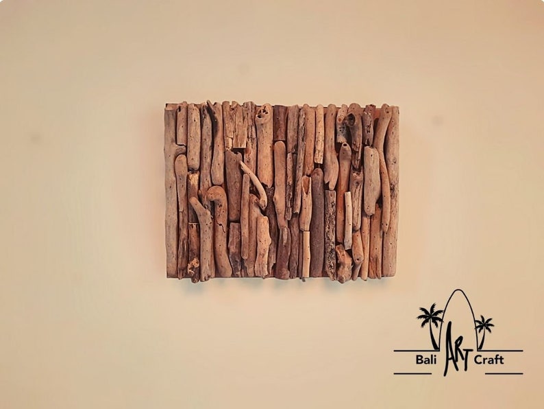 May include: A wall art piece made from driftwood, arranged in a vertical pattern. The wood is a light brown color and has a natural, rustic look. The piece is mounted on a white wall. The text "Bali Art Craft" is visible in the bottom right corner of the image.