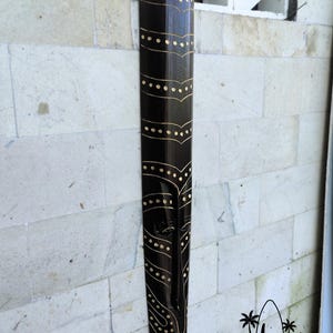 May include: A black wooden mask with a geometric design in gold paint. The mask is hanging on a white wall. Bali Art Craft.