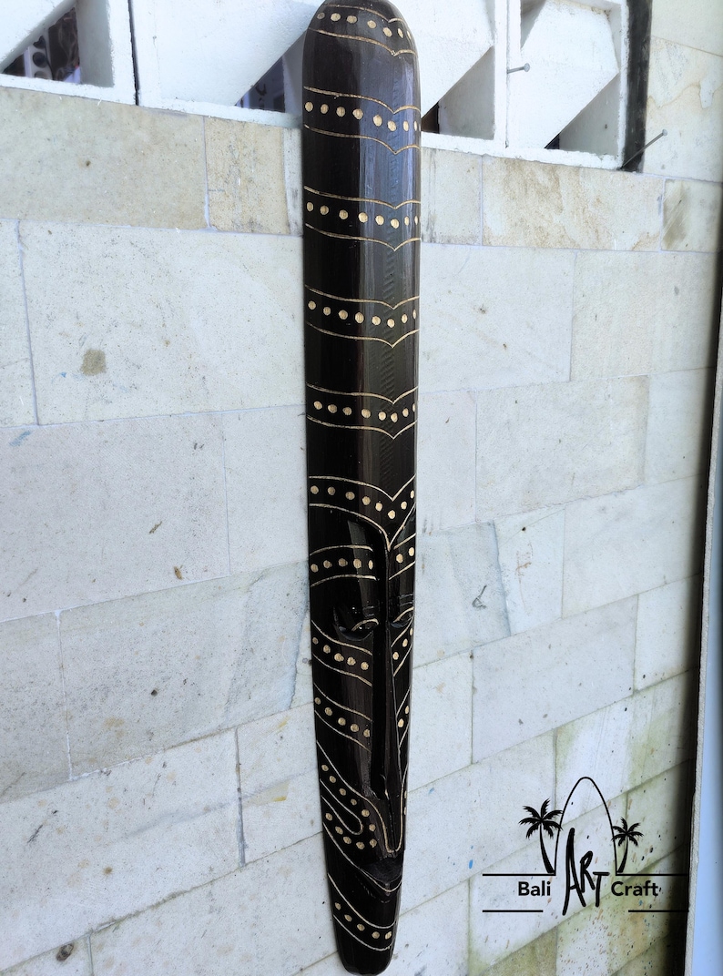 May include: A black wooden mask with a geometric design in gold paint. The mask is hanging on a white wall. Bali Art Craft.