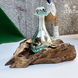 Op de afbeelding: Een heldere glazen fles met een ronde stop zit op een stuk drijfhout. De fles heeft een lichtgroene tint en is traanvormig. Het drijfhout is bruin en heeft een ruwe textuur.