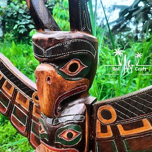 Puede incluir: Un tótem de madera tallado con un pájaro, pintado en tonos de marrón, verde, naranja y negro. El tótem tiene una cara estilizada con un pico grande y ojos. El tótem está decorado con patrones geométricos y puntos blancos.