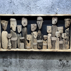 Op de afbeelding: Houten wandkunst met gesneden figuren met eenvoudige gezichten en gewikkelde details. De figuren zijn gerangschikt in een rechthoekige houten lijst tegen een donkere achtergrond. Het kunstwerk is bevestigd aan een getextureerde grijze muur.