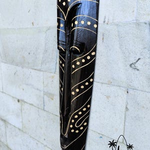 May include: Black wooden mask with a carved face and gold painted details. The mask has a geometric pattern of dots and lines. The mask is a traditional Balinese art form.