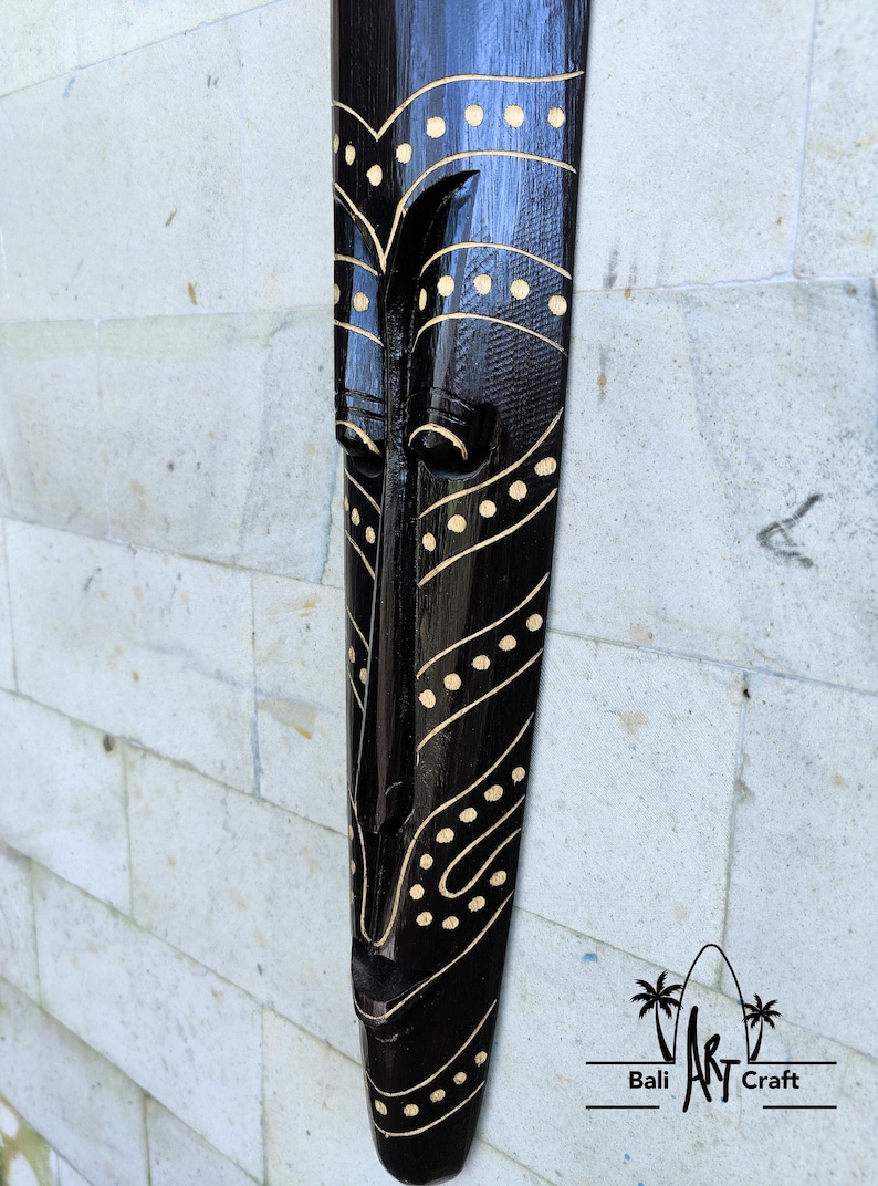 May include: Black wooden mask with a carved face and gold painted details. The mask has a geometric pattern of dots and lines. The mask is a traditional Balinese art form.