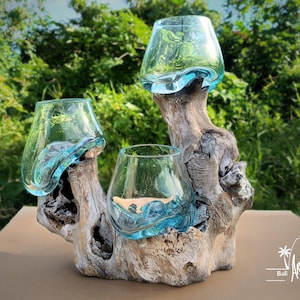 Può includere: Centrotavola decorativo con tre ciotole di vetro trasparente color turchese, ciascuna con un bordo unico a forma di onda, montate su una base di legno chiaro invecchiato. Il legno naturale ha un aspetto strutturato. Lo sfondo è un fogliame verde sfocato.