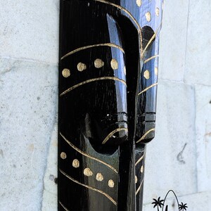 May include: Black wooden mask with gold painted details. The mask has a stylized face with a long nose and a wide mouth. The mask is decorated with a pattern of dots and lines.