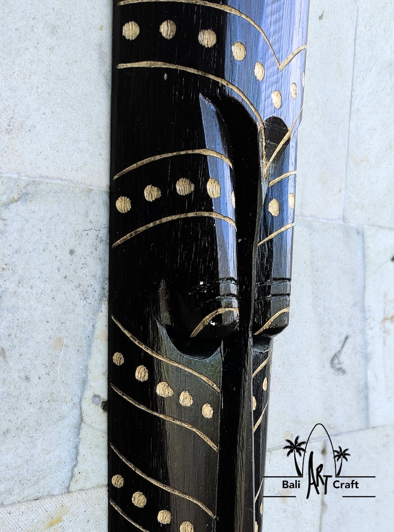 May include: Black wooden mask with gold painted details. The mask has a stylized face with a long nose and a wide mouth. The mask is decorated with a pattern of dots and lines.