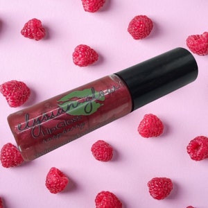 May include: A clear tube of red lip gloss with a black cap. The tube is labeled "Elysian Gloss Lip Gloss Raspberry". The tube is surrounded by red raspberries on a pink background.