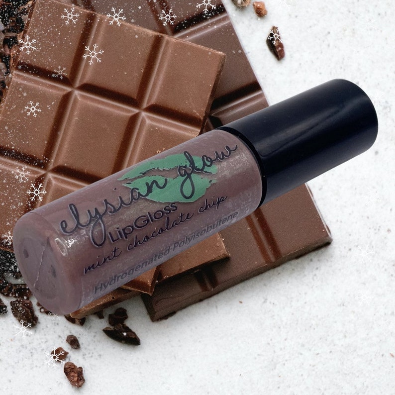 Puede incluir: Un tubo transparente con una tapa negra que contiene un brillo de labios marr&oacute;n. El tubo est&aacute; etiquetado con "Elysian Glow Lip Gloss mint chocolate chip Hydrogenated Polyisobutene". El tubo est&aacute; sobre una pila de barras de chocolate.