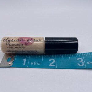 Puede incluir: Un peque&ntilde;o tubo transparente con una tapa negra que contiene un brillo de labios etiquetado como "elysian glow LipGloss cake batter" y "Hydrogenated Polyisobutene". El tubo se mide con una regla azul, midiendo aproximadamente 2,5 cm de largo.