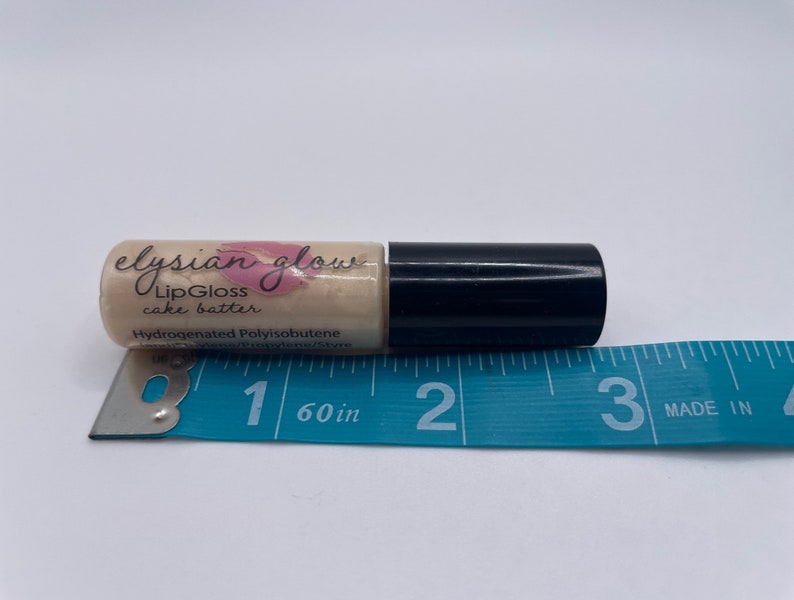 Puede incluir: Un peque&ntilde;o tubo transparente con una tapa negra que contiene un brillo de labios etiquetado como "elysian glow LipGloss cake batter" y "Hydrogenated Polyisobutene". El tubo se mide con una regla azul, midiendo aproximadamente 2,5 cm de largo.