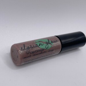 Puede incluir: Un tubo transparente con una tapa negra que contiene un brillo de labios marr&oacute;n claro. La etiqueta dice "elysian glow LipGloss mint chocolate chip".