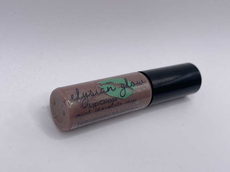 Puede incluir: Un tubo transparente con una tapa negra que contiene un brillo de labios marr&oacute;n claro. La etiqueta dice "elysian glow LipGloss mint chocolate chip".