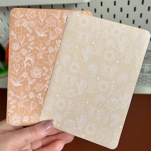 Peut inclure: Deux cahiers rectangulaires aux coins arrondis. L'un est de couleur pêche avec un motif floral et végétal blanc. L'autre est beige avec un motif blanc d'abeilles, de plantes et de fleurs. Les cahiers sont tenus dans une main.