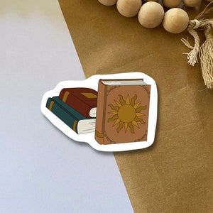 May include: A stack of three books with a sun design on the top book. The top book is brown with a gold sun design. The middle book is red and the bottom book is blue.
