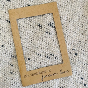 Op de afbeelding: Een bruin houten frame met een witte achtergrond en de tekst "it's that kind of forever love".