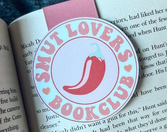 Könnte beinhalten: Ein runder Lesezeichen mit dem Text "SMUT LOVERS BOOK CLUB" in rosa Schrift. Eine rote Chili-Pfeffer-Grafik befindet sich in der Mitte. Das Lesezeichen liegt auf einem Buch mit einem rosa Tab.