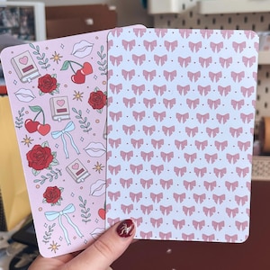 May include: Two rectangular cards. One card has a pink background with illustrations of roses, cherries, books, bows, and lips. The other card has a white background with a pattern of pink bows.