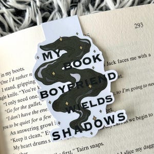 May include: A white bookmark featuring the text "MY BOOK BOYFRIEND WIELDS SHADOWS" in black. The design includes a dark green, swirling shape with gold star accents. The bookmark is in a book.