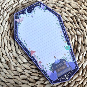 May include: A coffin-shaped notepad with light purple lined paper, featuring a dark purple border decorated with a crescent moon, stars, crystals, a moth, mushrooms, a potion bottle, and a stack of books labelled Spells, Potions, Magic & Mayhem.