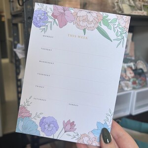 May include: A weekly planner notepad with pastel floral illustrations. The top of the notepad reads "THIS WEEK" in gold lettering. Days of the week are listed down the left side. Floral designs are in the corners.