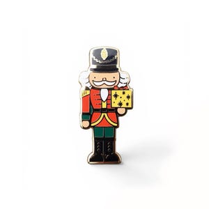 May include: A metal enamel pin shaped like a nutcracker soldier wearing a black hat, red jacket, and green trousers. The figure has white hair and a mustache, and is holding a yellow gift box decorated with black star patterns and a red bow.