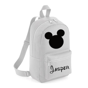 May include: A light grey backpack with a black Mickey Mouse silhouette. The bag has a front zipped pocket and the name "Jasper" printed in black script. It features adjustable shoulder straps and a top handle.