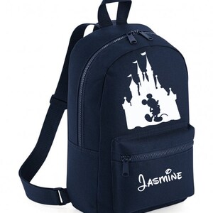 May include: A navy blue backpack featuring a white silhouette of Mickey Mouse in front of Cinderella's castle. The backpack has a zipped pocket on the front and the name "Jasmine" is printed below the image.