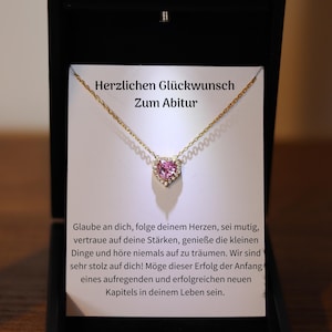May include: A gold necklace with a pink heart pendant set with diamonds. The necklace is in a black box with a white card that reads "Herzlichen Glückwunsch Zum Abitur" which translates to "Congratulations on your graduation".