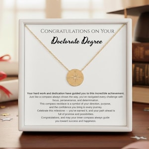 May include: A gold-colored compass necklace displayed in a white frame. The frame contains text that reads "CONGRATULATIONS ON YOUR Doctorate Degree." The necklace is on a chain. The background includes books and a graduation cap.