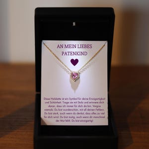 May include: A gold necklace with a pink heart pendant set with small diamonds. The necklace is in a white box with the text "An mein liebes Patenkind" on the front.
