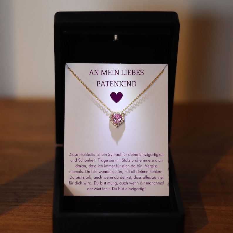 May include: A gold necklace with a pink heart pendant set with small diamonds. The necklace is in a white box with the text "An mein liebes Patenkind" on the front.