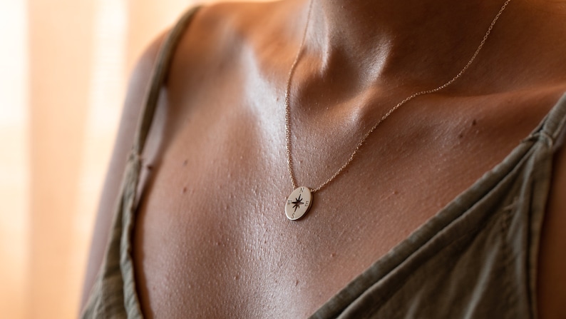 May include: A gold-colored pendant necklace featuring a compass design. The pendant is oval-shaped with a starburst pattern inside. The necklace is worn on a person's chest, with a green top visible.