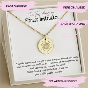 May include: A gold-colored necklace with a round pendant featuring a sunburst design. The necklace is presented in a white gift box with text that reads "To a Truly Amazing Fitness Instructor". The box also includes the text "Your dedication and strength inspire everyone around you every day."