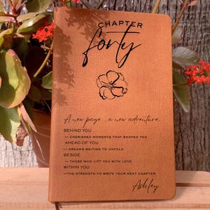 Peut inclure: Un journal en cuir marron avec l'inscription "Chapter Forty" en écriture élégante. Il présente une illustration florale et la phrase "A new page, a new adventure." Le journal contient également des citations inspirantes et le nom "Ashley."