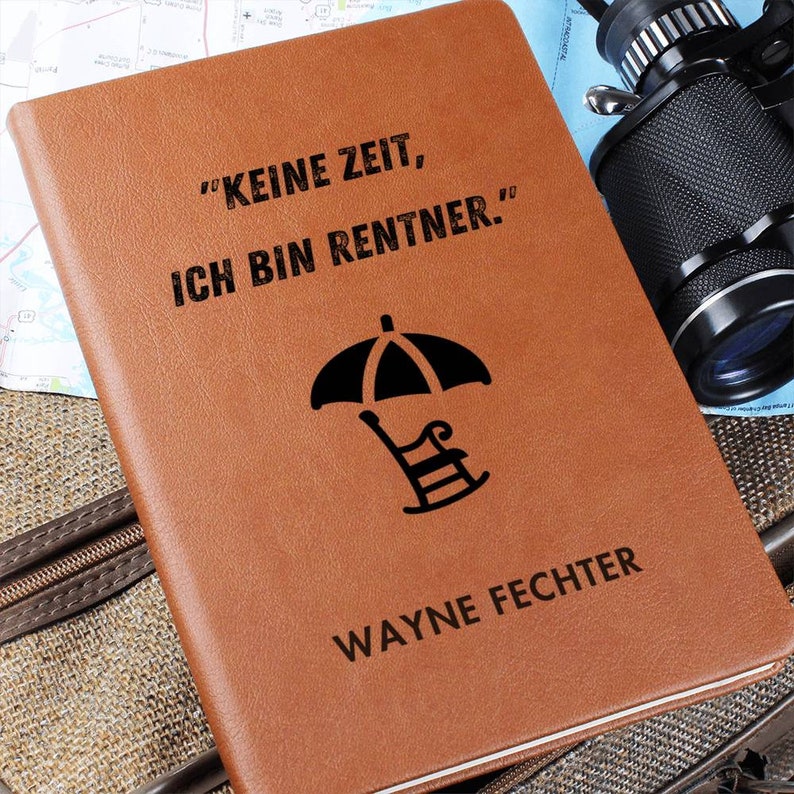 May include: A brown leather journal with the text "KEINE ZEIT, ICH BIN RENTNER." and a black silhouette of an umbrella and a chair. The name "WAYNE FECHTER" is printed below the image.
