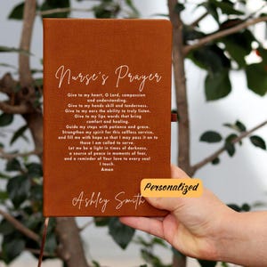 Può includere: Un diario in pelle marrone con le parole "Nurse's Prayer" incise sulla copertina. Il diario ha un'iscrizione bianca che recita "Give to my heart, O Lord, compassion and understanding. Give to my hands skill and tenderness. Give to my ears the ability to truly listen. Give to my lips words that bring comfort and healing. Guide my steps with patience and grace. Strengthen my spirit for this selfless service, and fill me with hope so that I may pass it on to those I am called to serve. Let me be a light in times of darkness, a source of peace in moments of fear, and a reminder of Your love to every soul I touch. Amen." Il diario è personalizzato con il nome "Ashley Smith".