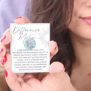 Könnte beinhalten: Eine weiße Karte mit dem Text "Difference Maker" und einer blauen und silbernen Anhänger-Halskette. Der Text auf der Karte lautet: "Ein engagierter Mensch, der die bemerkenswerte Fähigkeit besitzt, durch selbst kleinste Handlungen oder freundliche Worte einen bedeutenden Einfluss zu erzielen. Dies ist eine Person, die das Leben anderer konsequent verbessert, als Inspiration dient, die sie vorwärts treibt und positive Veränderungen bewirkt."