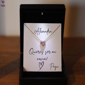 May include: A gold necklace with a heart-shaped pink gemstone pendant, presented in a black jewelry box. The box contains a card with the name "Alexandra" and the Spanish text "Quieres ser mi novia?" with a heart and the name "Paqui".
