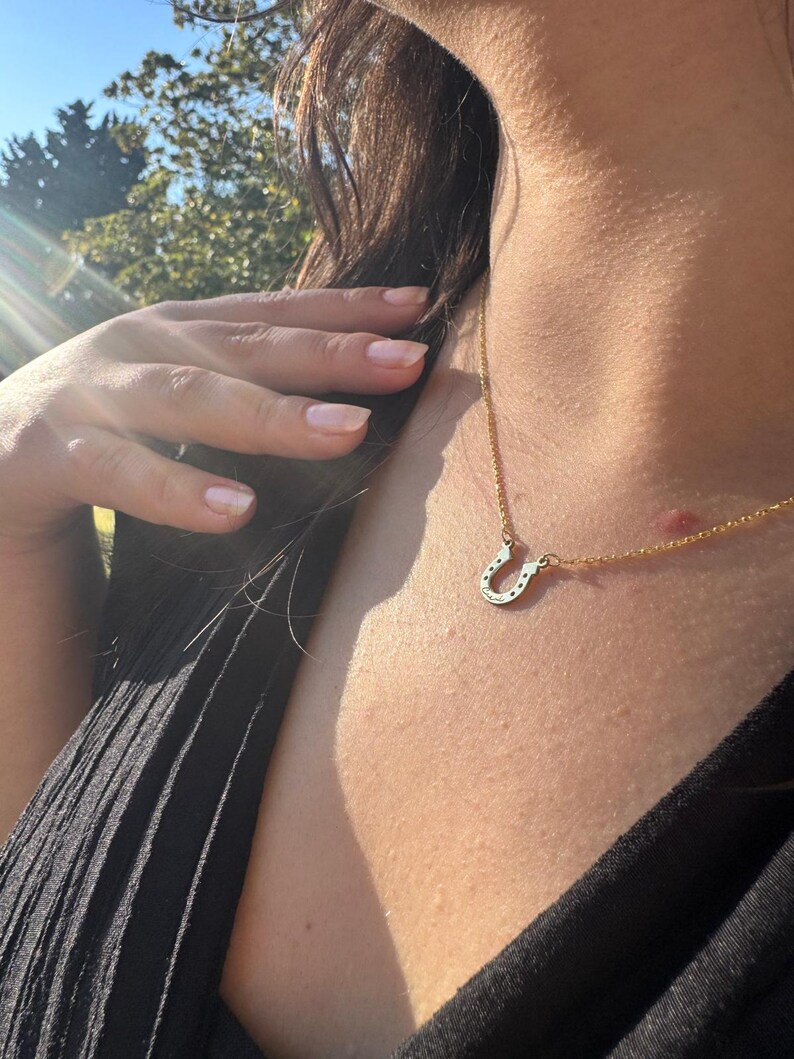May include: A gold-colored necklace with a horseshoe pendant. The horseshoe is detailed with small, sparkling accents. The necklace is worn against a black top, with a hand gently touching the hair. The background is blurred with sunlight.
