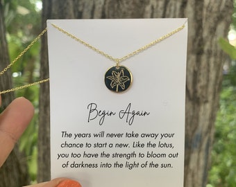 May include: Gold necklace with a black pendant featuring a gold lily design. The pendant is on a white card with the text "Begin Again" and a quote about the strength to bloom out of darkness.